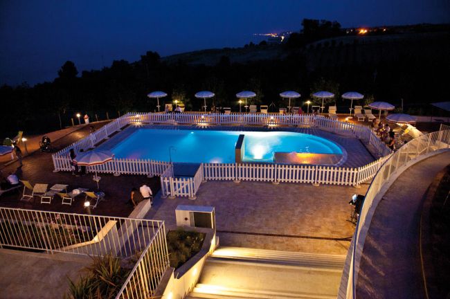 Piscina illuminata di notte, circondata da recinzione bianca e sdraio con ombrelloni.