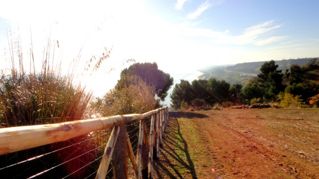 Sentiero sterrato con recinzione in legno, vista panoramica su colline e mare al tramonto.