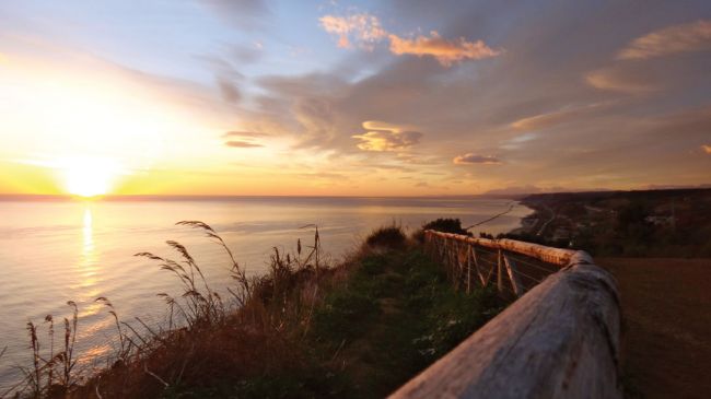 Un tramonto su una costa vista da un sentiero con staccionata.