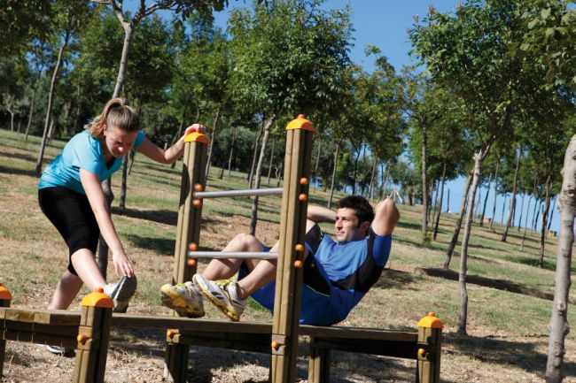 Due persone si allenano su attrezzature all'aria aperta in un parco alberato.