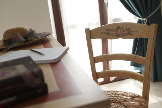 Una scrivania con sedia decorata, un quaderno aperto e un cappello di paglia vicino a una finestra.