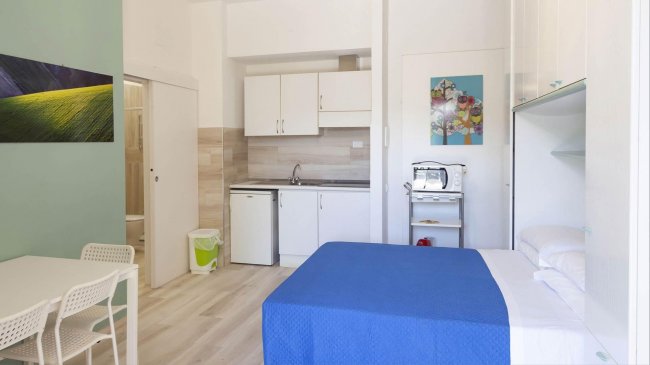 Camera con angolo cottura, letto blu e tavolo bianco.