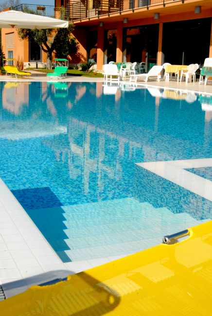 Dettaglio della piscina con scalini bianchi, riflessi nitidi e lettini gialli, immersa nel sole.